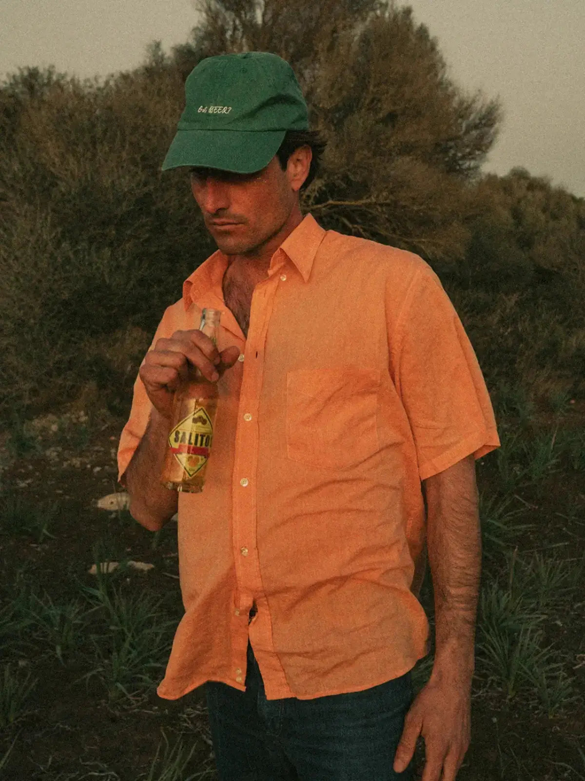 Ein Mann steht draußen, hinter ihm sind Bäume, er trägt ein orangenes Hemd und eine grüne SALITOS Cap.