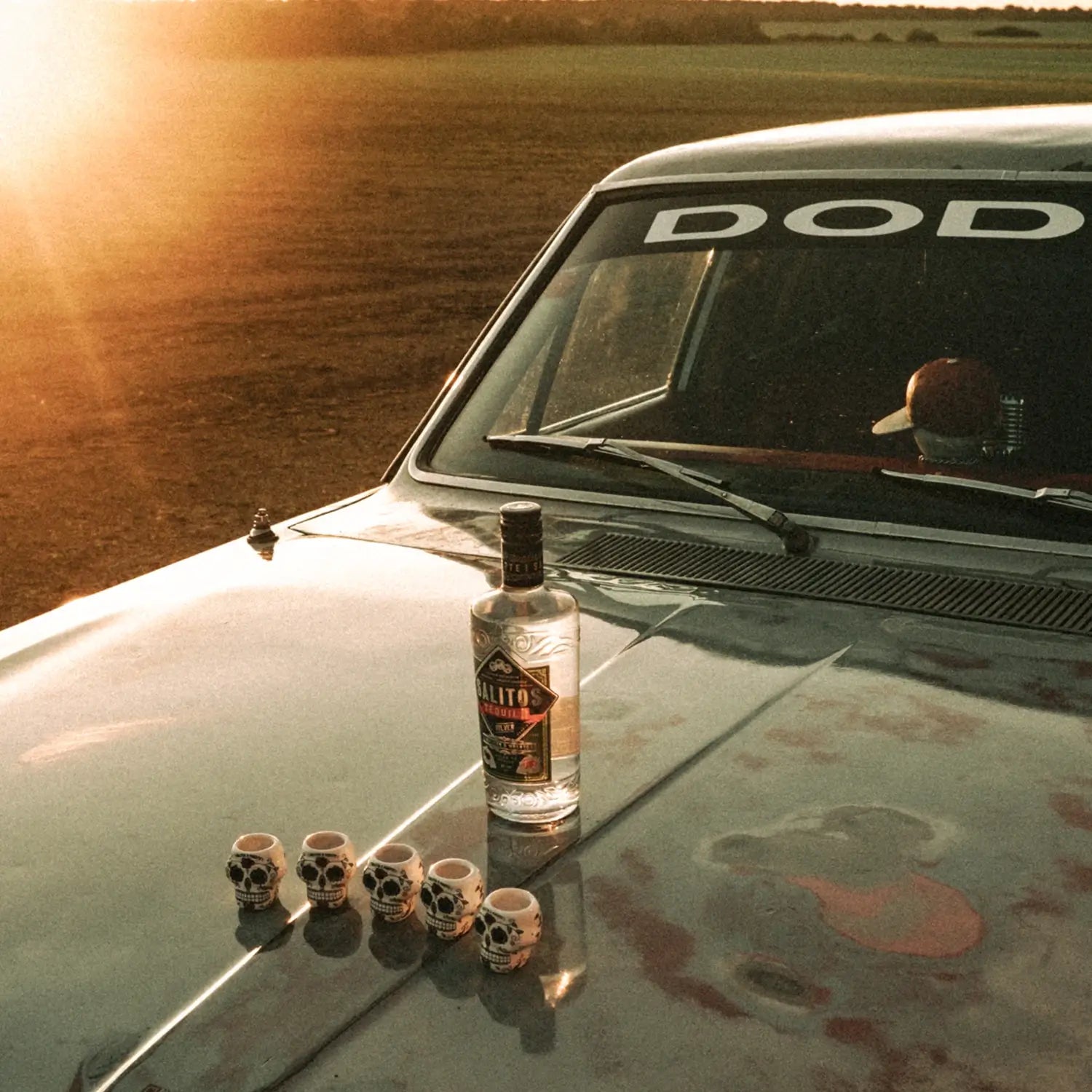 Ein Auto welches auf einem Feld in der Sonne steht. Auf der Motorhaube befinden sich 5 Shot-Gläser die aussehen wie Totenköpfe, sowie eine Flasche SALITOS Tequila Silver