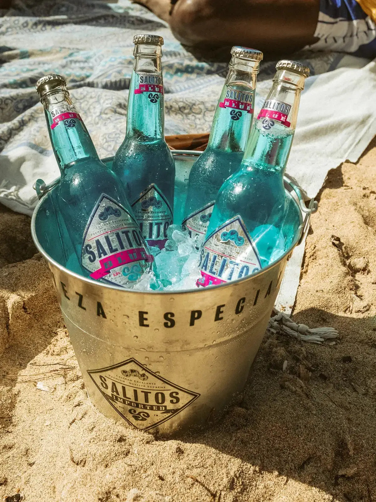 Ein Metalleimer der auf Sand steht und gefüllt mit SALITOS Blue Flaschen, sowie Eiswürfeln ist.