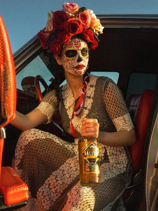 Eine Frau geschminkt wie ein Totenkopf hat eine Krone aus Blumen und in der Hand hält Sie eine Flasche SALITOS Tequila Gold