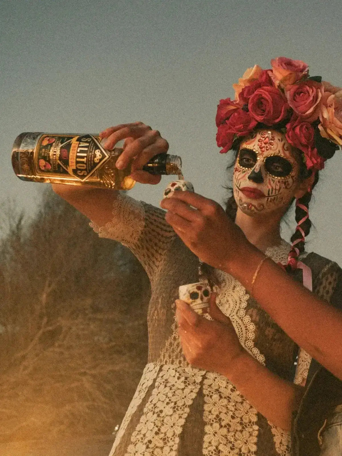 Eine Frau geschminkt wie ein Totenkopf hat eine Krone aus Blumen und in der Hand hält Sie eine Flasche SALITOS Tequila Gold und schenkt dies in Gläser ein.